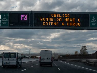 Araçlar bulutlu bir gökyüzünün altında, üzerinde elektronik bir yol tabelası olan bir otoyolda İtalyanca 'da 