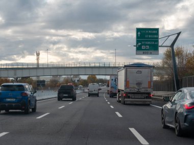 Araclar bulutlu bir gökyüzünün altında arka planda bir köprü, Milano, Venezia ve Belluno 'yu gösteren yön işaretleriyle dolu yoğun bir otoyolda ilerliyorlar. Vicenza, İtalya - 21 Kasım 2025