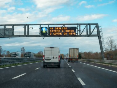 Araçlar, bulutlu bir günde seyahat bilgilerini gösteren elektronik bir yol tabelasının altında otoyolda araba kullanırlar. Vicenza, İtalya - 21 Kasım 2025