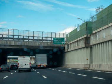 Araçlar yoğun bir otoyolda üstgeçitle ve yön tabelasıyla, dağınık bulutlu açık mavi bir gökyüzüne doğru ilerliyorlar. Vicenza, İtalya - 21 Kasım 2025