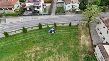 Bir yerleşim bölgesinde traktörle çim biçen bir çiftçinin hava görüntüsü, kentsel ve kırsal yaşamın birlikte yaşadığını ve bir kasaba içindeki yeşil alanların bakımını gösteriyor.