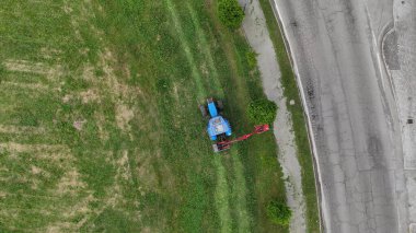 Yol boyunca traktör ve çim biçme makinesiyle çim biçen bir çiftçinin hava görüntüsü, iyi korunmuş ve resimli kırsal bir manzaraya katkıda bulunuyor.