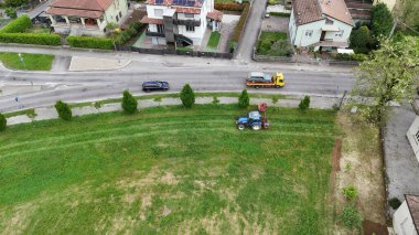 Yerleşim bölgesinde bir traktörle çim biçen çiftçi, bir çekici araç yol boyunca bir araba taşırken ve başka bir araç geçerken, canlı bir kırsal alan yaratıyor.