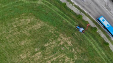 Bir çiftçinin traktörle çimenleri biçtiği hava görüntüsü, yoldan geçen bir otobüsün yanında, kırsal ve kentsel yaşamın kesişimlerini gösteriyor.