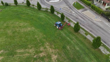 Tarım traktörü titizlikle aşırı büyümüş otları ve uzun otları kalabalık bir kasaba yoluna bitişik halka açık yeşil alandan temizliyor ve şehir estetiğini geliştiriyor.