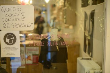 İtalyan mağaza vitrininde COVID 19 'un maske gerekliliği ve maksimum kapasiteyle ilgili güvenlik işaretleri görülüyor. Yanında bir manken ve camda şairane bir yazı var. Hepsi sokaktan gelen sahte ışıkları yansıtıyor.