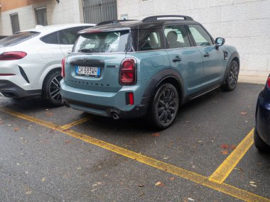 Modern mavi bir Mini Cooper vatandaşı, sarı çizgileri olan ıslak bir asfalt yüzeyinde belirlenmiş bir yere park etti.