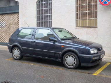 Klasik bir koyu mavi üç kapılı Volkswagen Golf Mk3 hatchback şehir merkezinde sarı çizgilerle işaretlenmiş asfalt bir yüzeye park edilmiştir.