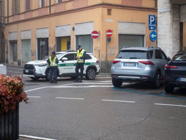 Üniformalı iki polis memuru, arka planda park etmiş araçlar ve binalar olan bir caddede devriye arabasının yanında dururlar.