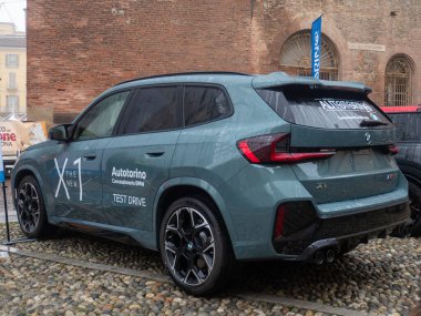Modern bir BMW X1 SUV, tanıtım amaçlı bir test sürüşü etkinliği sırasında sergilenmektedir, tasarımını ve özelliklerini eski mimarinin bir arka planına karşı vurgulamaktadır.