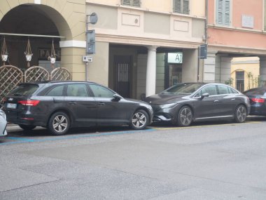 İki modern sedan, bir Audi A4 Avant ve bir Mercedes-Benz EQE, arka planda binaları olan nemli bir şehir yolunda yan yana park edilmişlerdir.