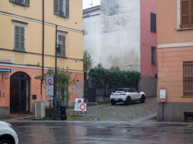 Modern beyaz bir elektrikli arazi aracı bulutlu bir günde renkli binalar arasında ıslak bir kaldırım taşından ara sokağa park edilmiştir.