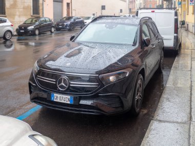Parlak siyah bir Mercedes-Benz EQA elektrikli SUV ıslak, yansıtıcı bir caddeye park edilmiş ve yüzeyinde yağmur damlaları var.