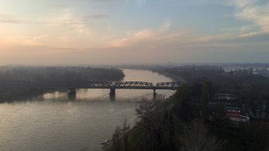 Geniş, sakin bir Po Nehri, sisli bir manzarası ve alacakaranlıkta görünen uzak şehir siluetiyle tarihi bir demiryolu köprüsünün altından akar.