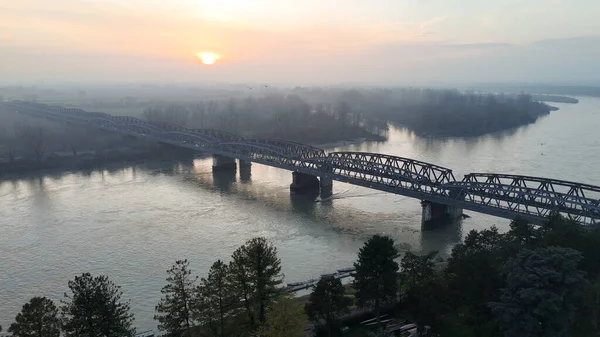 Güneş sisli bir gökyüzünden yükseliyor, Po Nehri 'nin üzerine yumuşak bir parıltı saçıyor ve Cremona, Lombardy' deki tarihi demiryolu köprüsü