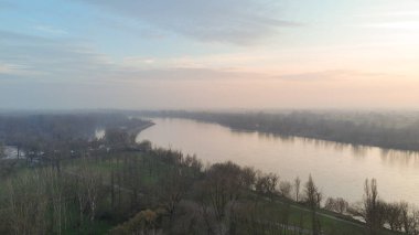 Geniş bir nehir, şafak vakti yumuşak, sisli bir gökyüzünün altındaki ağaçların ve tarlaların arasından akar ve İtalyan kırsalının huzurlu güzelliğini gözler önüne serer.