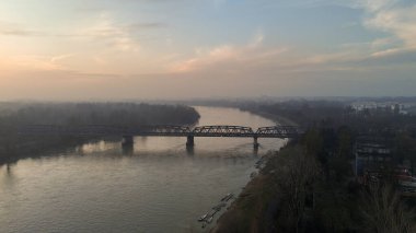 Geniş bir hava perspektifi, İtalya 'nın Cremona kentinden akan Po Nehri' ni, sularını sisli bir günbatımı gökyüzü altında kaplayan tarihi bir demiryolu köprüsü ile yakalıyor.