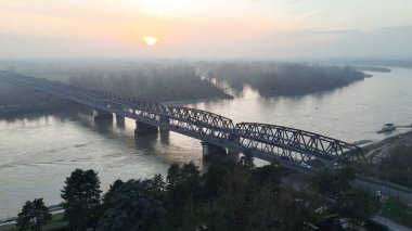 Güneş puslu bir gökyüzünün ardında batıyor, Po Nehri 'nin üzerine sıcak bir parıltı saçıyor ve Cremona, Lombardy' deki tarihi demiryolu köprüsü