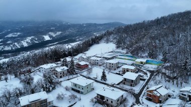 Taze karla kaplı, bulutlu bir gökyüzü altında orman ve tepelerle kaplı küçük bir İtalyan dağ köyünü gösteren sakin bir kış manzarası Le Bore, PR, İtalya