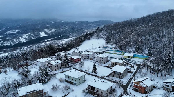 Taze karla kaplı, bulutlu bir gökyüzü altında orman ve tepelerle kaplı küçük bir İtalyan dağ köyünü gösteren sakin bir kış manzarası Le Bore, PR, İtalya