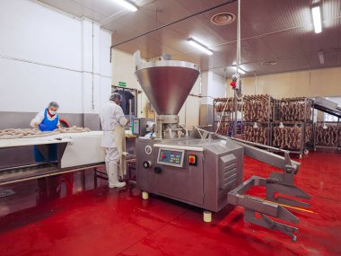 Temiz et işleme tesisinde çalışan işçiler, sosis doldurma makineleri kullanırlar ve pişirilmiş etleri üretim için raflara asarlar.