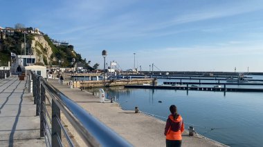 Numana, İtalya - 22 Nisan 2023 Güneşli bir günde sakin sulara yanaşmış liman ve teknelerin manzarasını seyreden bir kadın, sakin bir rıhtımda sakin bir yürüyüş yapıyor..