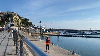 Numana, İtalya - 22 Nisan 2023 Rıhtımda tek başına duran sakin ve huzurlu bir kadın sahnesi, sakin denizi ve açık mavi gökyüzünü alıyor. Rahatlamak için mükemmel bir ortam.