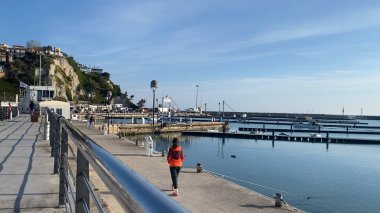 Numana, İtalya - 22 Nisan 2023 Güneşli bir günde sakin bir deniz ve berrak mavi gökyüzü, seyahat ve turizm reklamı için mükemmel bir deniz kenarında sakin bir yürüyüş yapan bir insan.,