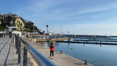 Numana, İtalya - 22 Nisan 2023 turuncu ceketli bir kadın huzurlu bir rıhtım boyunca yürüyor, sakin atmosferi ve liman ve mavi gökyüzünün çarpıcı manzarasını seyrediyor. Mükemmel.