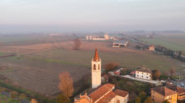 İtalya 'nın Cremona kentindeki Vescovato kentindeki San Bartolomeo köyünü kapsayan hava aracı görüntüsünde, geniş tarım alanları ve gün batımında ana yol bulunan kilise ve çan kulesi yer alıyor.