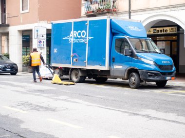 Cremona, İtalya - 24 Aralık 2025 Mavi bir teslimat kamyonu, güvenlik yeleği giymiş bir işçiyle birlikte şehir caddesinde park etmiş sarı süpürgeyle yolu temizliyor..