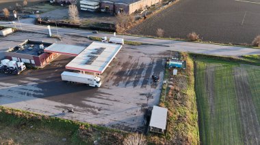 Doğal ışık altında tarım arazileri ve sanayi yapılarının yanına kurulmuş büyük kamyon ve arabaların yer aldığı bir taşra otoyolu üzerindeki bir benzin istasyonu ve servis alanının genel görünümü