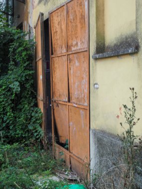 Paslı bir kapısı olan terk edilmiş bir bina aşırı gelişmiş bitki örtüsüyle çevrili. Terkedilmiş ve ihmal edilmiş bir hissi uyandırıyor..
