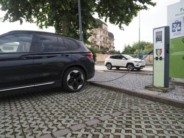 Crema, İtalya - 5 Temmuz 2025 Yakınlarda beyaz bir SUV ile bir şehir istasyonunda siyah elektrikli araba şarj ediliyor, modern bir şehirde sürdürülebilir ulaşım sergileniyor..