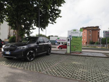 Crema, İtalya - 5 Temmuz 2025 Modern bir kentsel şarj istasyonunda parlak siyah bir BMW elektrikli araba, şehir binaları, ağaçlar ve bulutlu bir gökyüzü ile çevrili, sürdürülebilir enerji ve