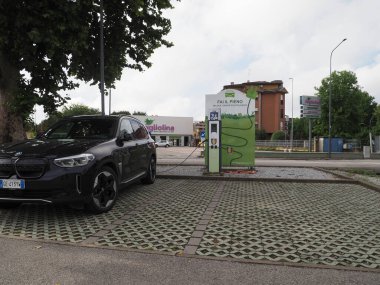 Crema, İtalya - 5 Temmuz 2025 Modern yeşil şarj istasyonunda parlak siyah bir BMW elektrikli araba, bulutlu bir gökyüzünün altındaki ağaçlar ve binalarla çevrili,