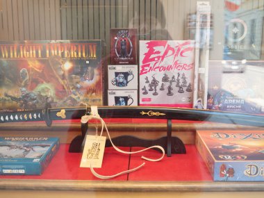 Cremona, İtalya 9 Ocak 2026. Dükkan vitrininde bir katana, Twilight Imperium, Warhammer, Dixit ve Sorcerer 's Arena gibi masa oyunları, artı kupalar ve minyatürler sergileniyor.