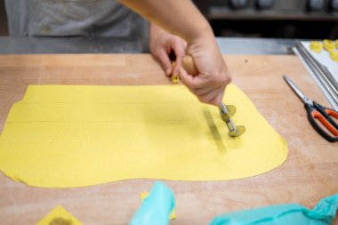 Şefin elleri, ahşap bir yüzey üzerinde hamur işi tekerleği ile şeritlere taze sarı makarna hamuru keserek, profesyonel bir mutfakta otantik İtalyan yemekleri için geleneksel tortelli hazırlıyor.