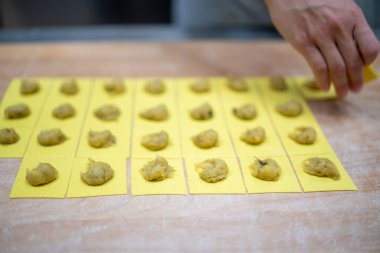 Şefin elleri ahşap bir tahtanın üzerinde taze el yapımı tortelli dizileri oluşturuyor. Sarı hamur karelerini profesyonel bir İtalyan mutfağında lezzetli malzemelerle dolduruyor.