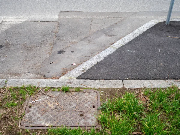 Yıpranmış, elmas desenli metal bir kapak beton kaldırımı ve asfaltı olan bir caddenin yanında yeşil çimlerle kısmen gizlenmiş duruyor.