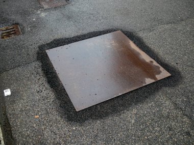 Asfaltı kaplayan ıslak metal plaka. Yakındaki ızgara ve döküntülerle birlikte.