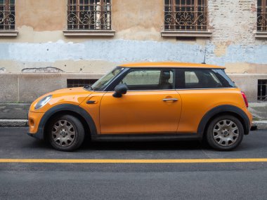 Cremona, İtalya 6 Şubat 2026 Orange Mini Cooper hatchback sarı çizgi işaretli asfalt sokağa park etmiş, arka planda sıkıntılı bir bina cephesi