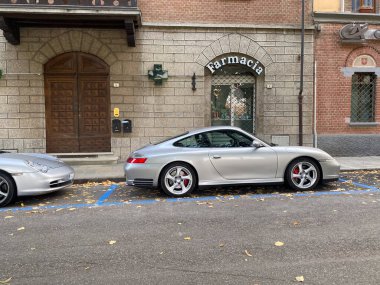 Bir asfalt yolda yan yana park etmiş iki gümüş renkli Porsche 'nin yer aldığı bir sokak sahnesi.