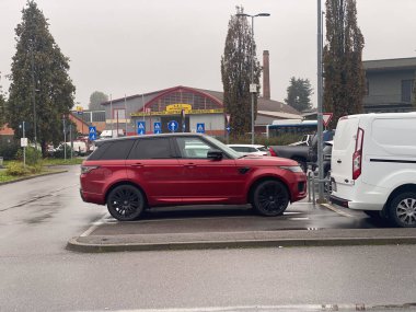 Modern kırmızı Range Rover spor bir SUV, yanında beyaz bir minibüs bulunan bir otomobil tamirhanesinin yakınında park halinde.