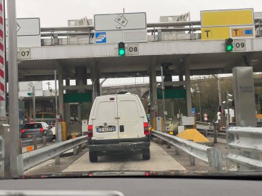 Kasvetli bir bulutlu günde, birden fazla şeridi açık ve trafiği tıkalı beyaz bir minibüs gişeye yaklaşıyor.