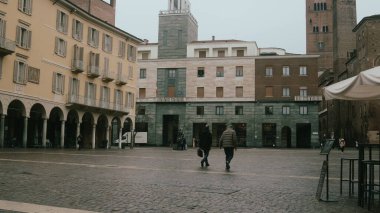 Cremona, İtalya. 6 Şubat 2026. Piazza del Comune, Cremona, Lombardy, İtalya. Bulutlu bir kış gününde tarihi mimarisi ve yürüyen iki figürle.