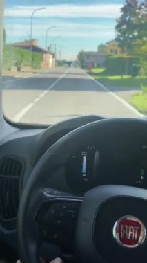 POV bir arabanın içinden çekilmiş, direksiyonu ve gösterge panelini gösteriyor, önünde kırsal bir yol ve binalar var.