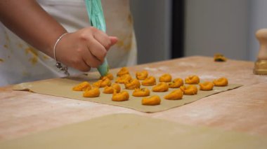 Parşömen kağıdının üzerinde parlak, temiz bir mutfak ortamında küçük, turuncu tortelli makarna şekilleri oluşturmak için boru torbası kullanan bir kadının ellerine yakın çekim.
