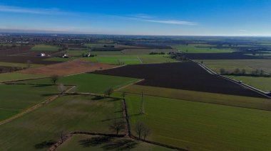 Uçsuz bucaksız Padana ovasında Po Vadisi tarım arazileri: güneşli bir öğleden sonra açık mavi gökyüzü altında yeşil ekinler ve tarım binaları, ağaçlar ve bir kır yolu olan karanlık toprak parçaları