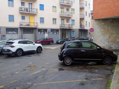 Cremona, İtalya - 3 Şubat 2026 Siyah bir Lancia Ypsilon ve beyaz bir Kia Sportage zemin katta dükkanları olan apartmanların önündeki ıslak asfalt bir caddede park halindeler.